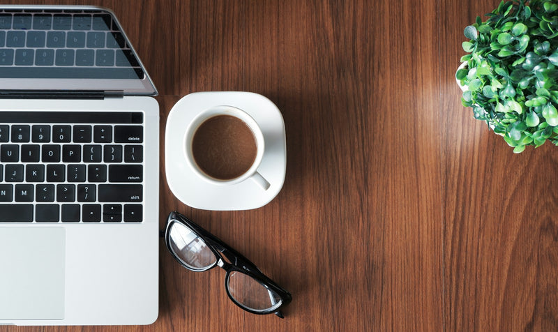 Overhead view of a warm walnut home-office desk with a laptop, coffee cup, glasses and a small plant